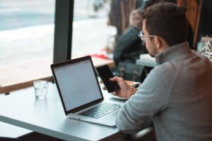 Person holding phone to research Interfax vs Etherfax comparisons while sitting at a table with their laptop